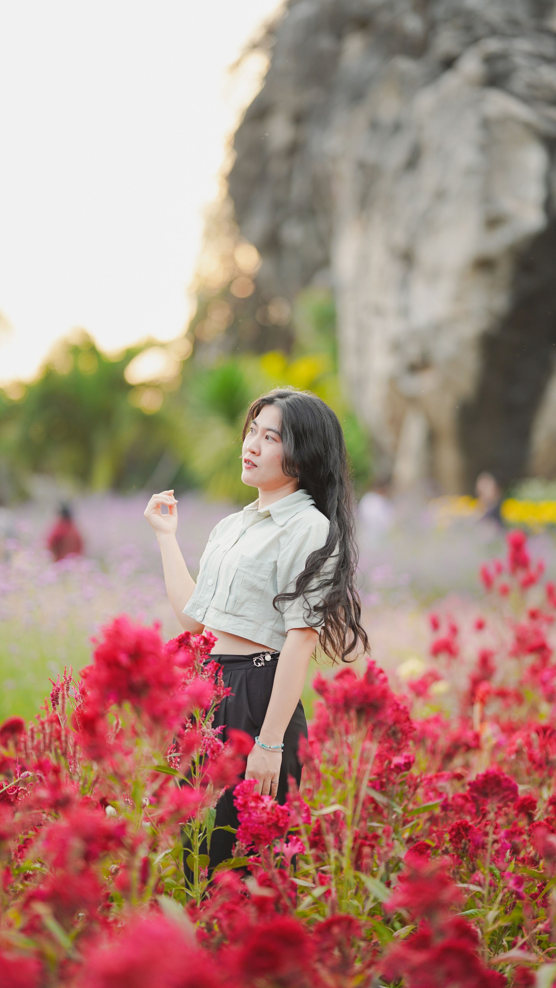 A young woman stands amidst vibrant red flowers, with a natural landscape in the background, showcasing beauty and tranquility.