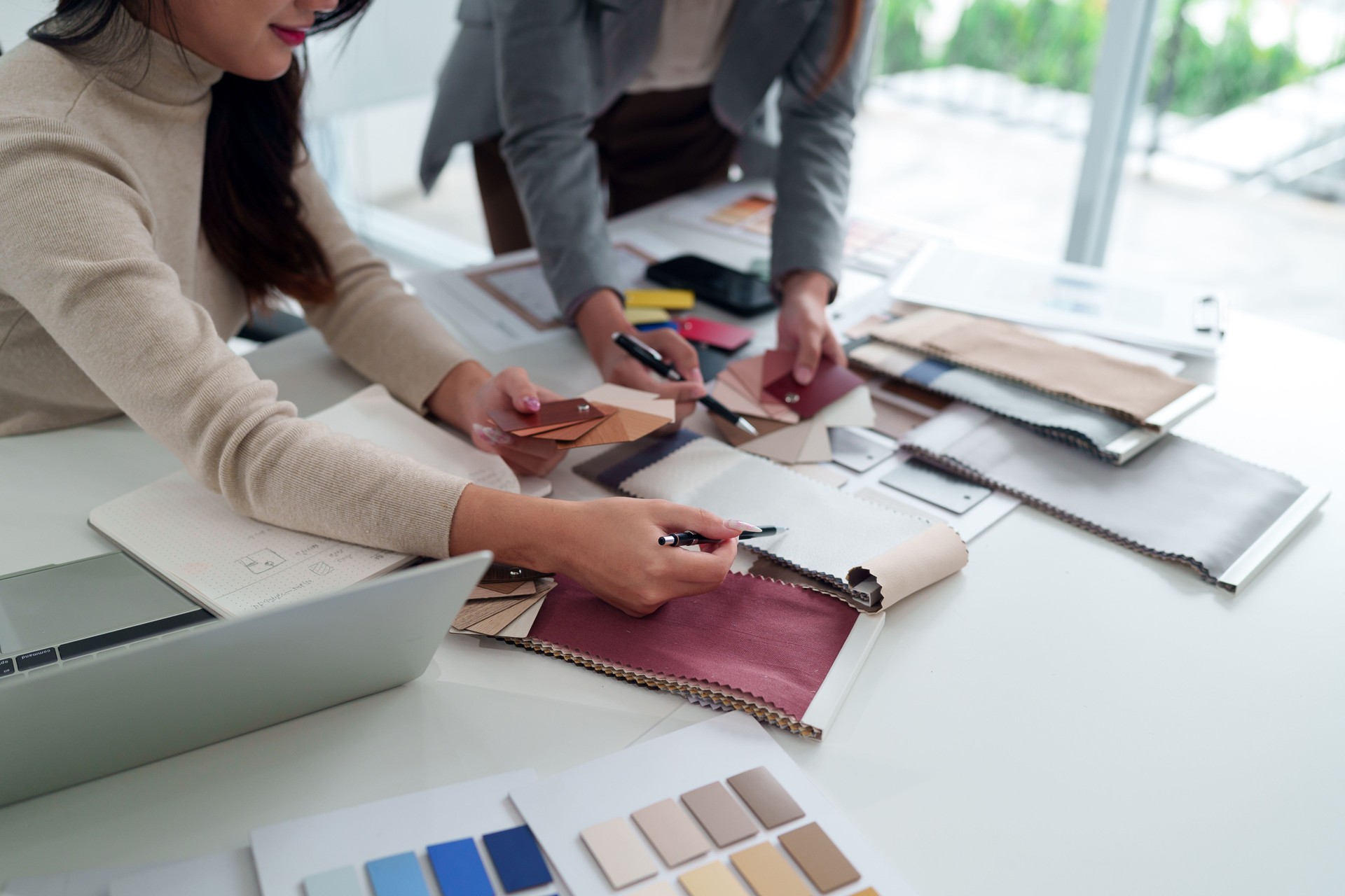 Asian Women Interior Designers Collaborate on Fabric Selection for Home Decor, Planning Design Project with Color Swatches and Textile Samples in Studio Space